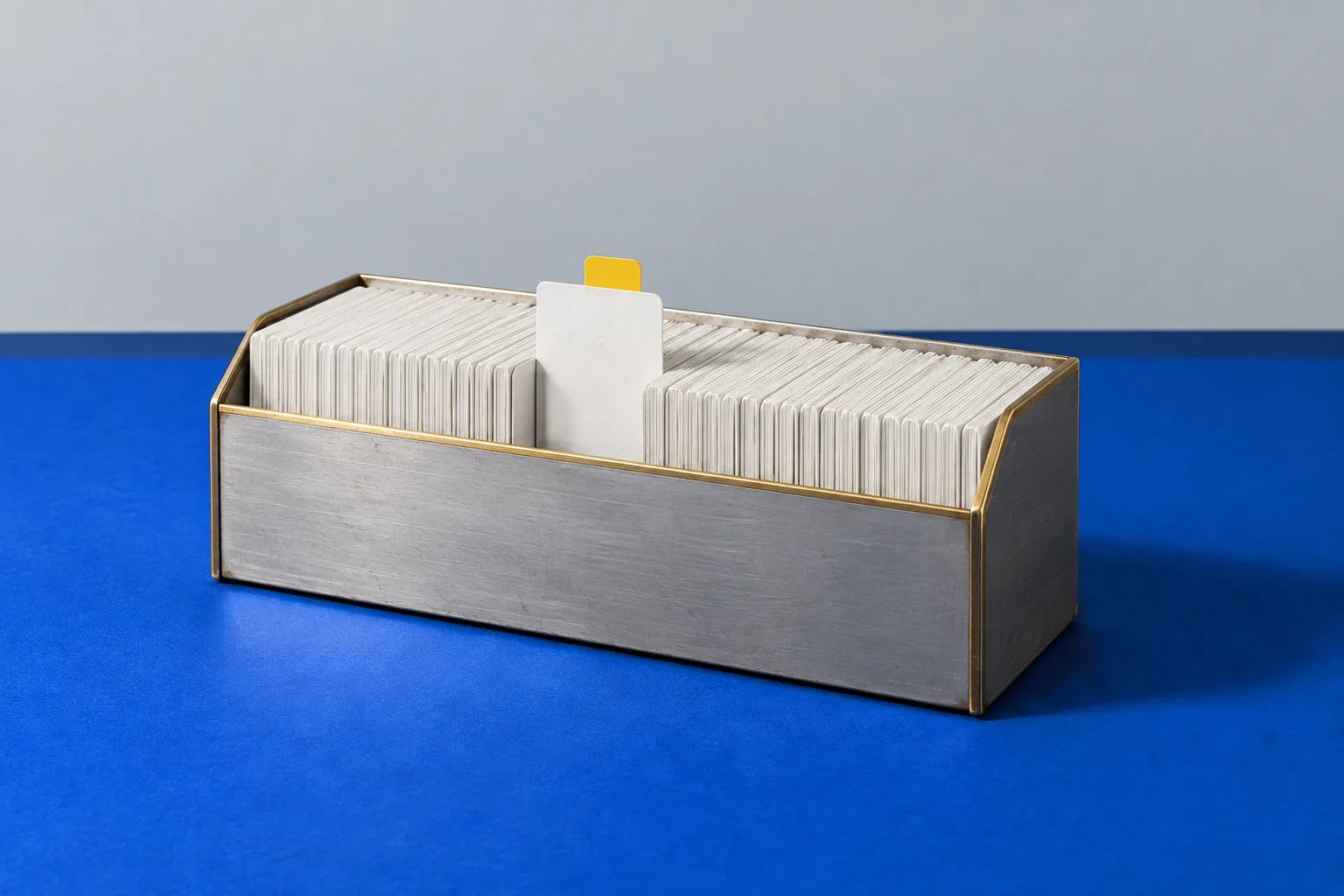 A brushed-metal card box full of white cards, one with a yellow tab, on a blue floor against a light wall.